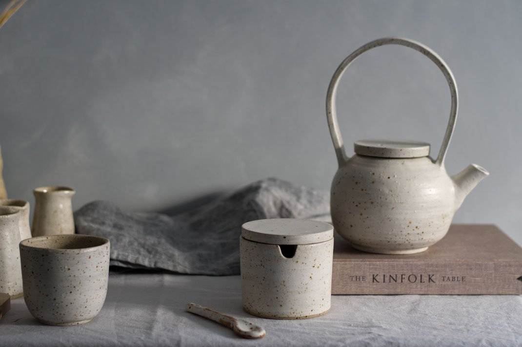 Verschillende stukken handgemaakt keramiek van gespikkelde klei waardonder een suikerpotje, een kopje en een theepot op een boek geplaatst