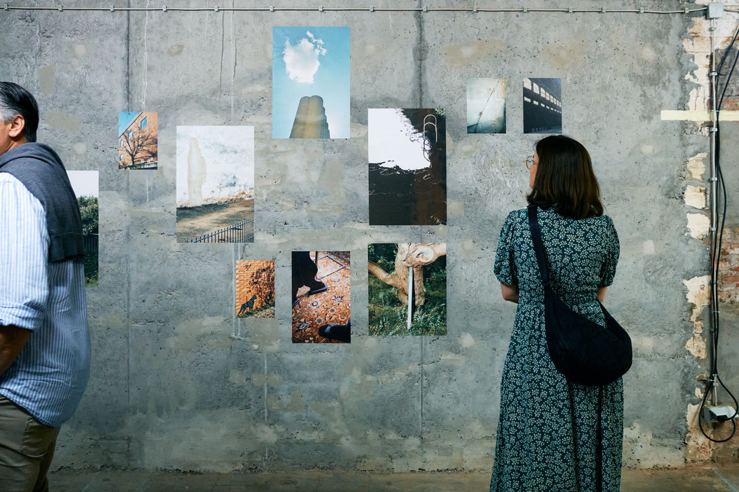 Eine Person in ornametiertem Kleid und schwarzer Umhängetasche schaut sich Ausstellungsbilder der Ausstellung "wegsehen zwecklos" an. Die Wand, auf der die Bilder aufgeklebt sind, ist halb-glattgestrichener, grauer Beton.