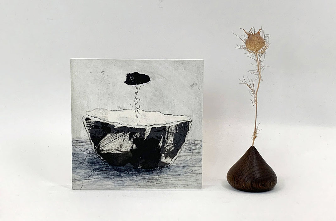 Schwarz-weißes Yūgen-Wandobjekt neben einer minimalistischen Holzvase mit einer vertrockneten Blütenkapsel. Stillleben-Arrangement im Wabi-Sabi-Stil.