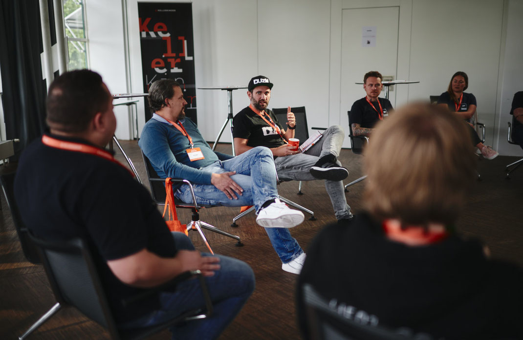 Event Foto vom BAR-CAMP 2022: Teilnehmer sitzen im Stuhlkreis, ein Mann mit schwarzer DUZN-Mütze spricht und gestikuliert, im Hintergrund ist ein Roll-Up mit dem Schriftzug „Kellerkinder“ zu sehen.