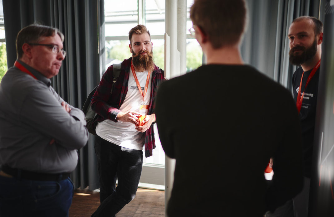 Event Foto vom BAR-CAMP 2022: Fünf Männer stehen in einer lockeren Gesprächsrunde am Fenster, teils mit Namensschildern und orangenen Lanyards, im Hintergrund dunkle Vorhänge und Tageslicht.
