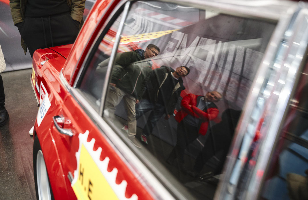 Event Fotos mit Spiegelung: In der Lackierung des legendären AMG-Rennwagens spiegeln sich drei Teilnehmer des Workshops – ein kreatives Beispiel dafür, wie Event Fotos auch ungeplante Kompositionen und Markenidentität einfangen können.