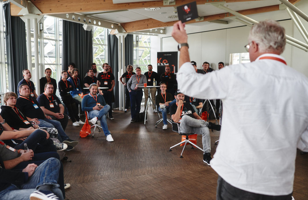 Event Foto vom BAR-CAMP 2022 der KELLERKINDER: Eine größere Gruppe von Teilnehmenden sitzt und steht in einem hellen Raum, während ein Mann im Vordergrund eine Karte hochhält und spricht.