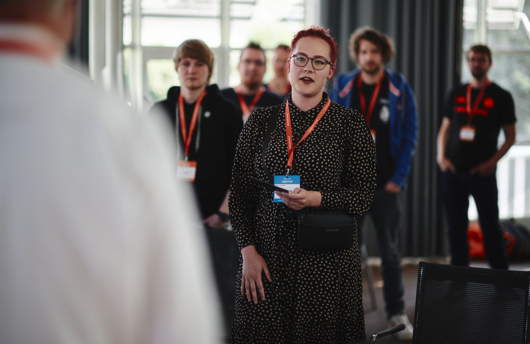 Event Foto vom BAR-CAMP 2022 der KELLERKINDER: Eine Teilnehmerin mit gepunktetem Kleid und rotem Haar spricht vor einer Gruppe, mehrere Zuhörer stehen im Hintergrund.