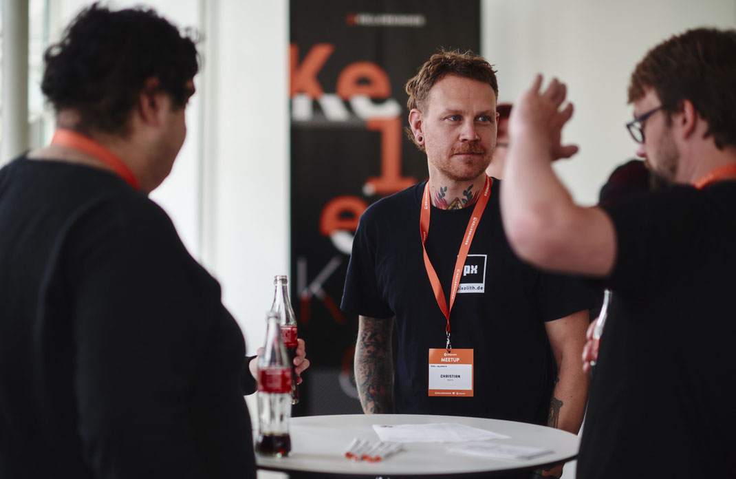 Event Foto vom BAR-CAMP 2022 der KELLERKINDER: Drei Männer stehen an einem Stehtisch, unterhalten sich und trinken Cola. Im Hintergrund ist ein Banner mit dem Schriftzug „KELLERKINDER“ zu sehen.