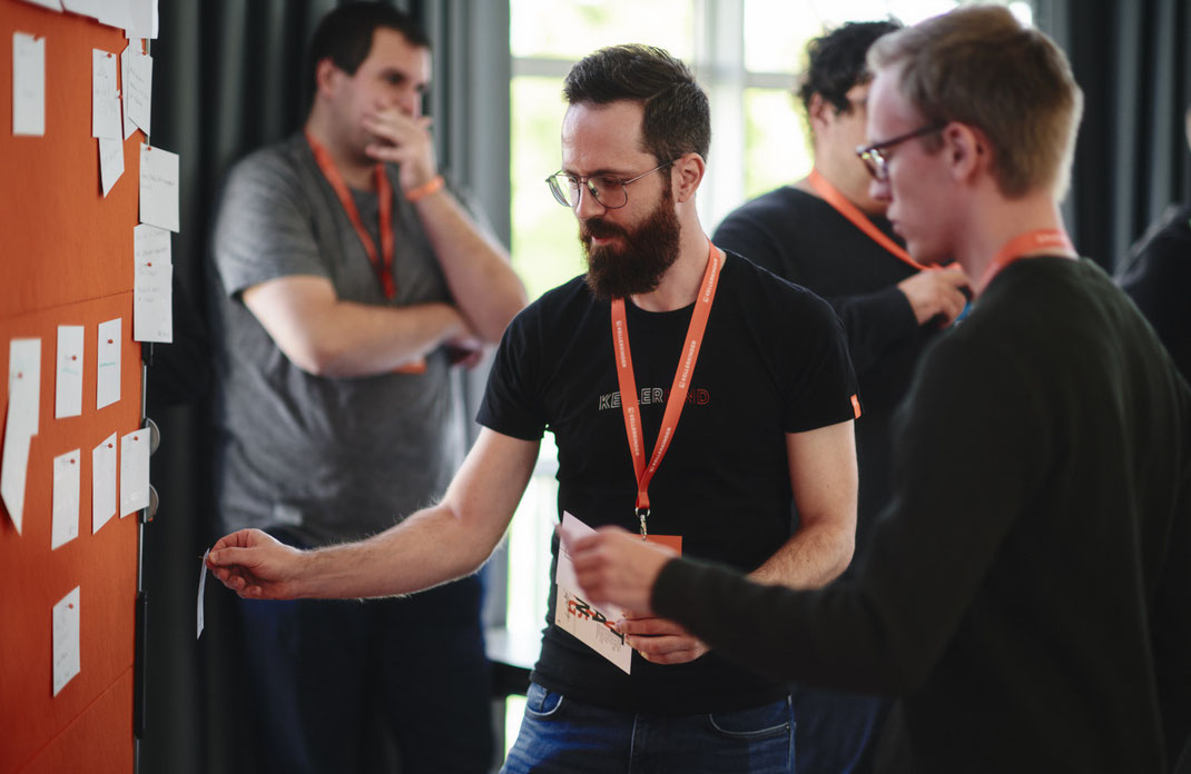 Event Foto vom BAR-CAMP 2022 der KELLERKINDER: Mehrere Teilnehmende stehen vor einer orangefarbenen Pinnwand mit vielen Notizzetteln, einer davon pinnt gerade einen weiteren Zettel dazu.
