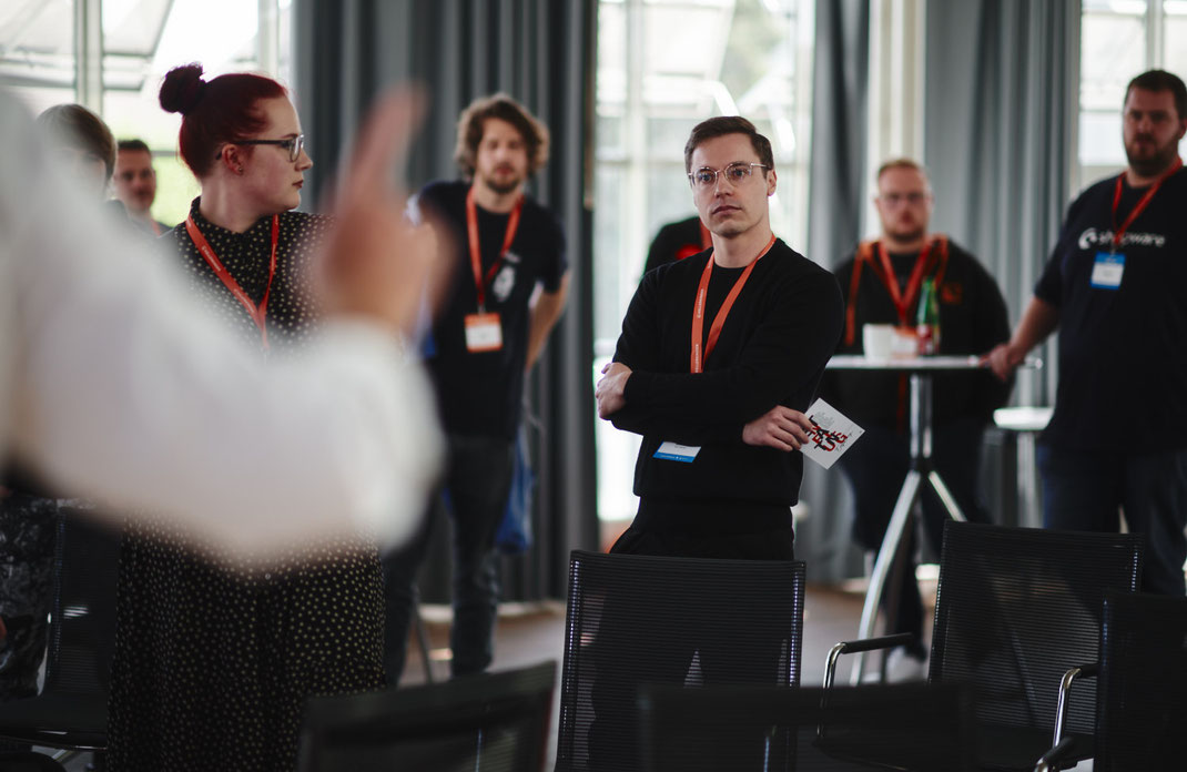 Event Foto vom BAR-CAMP 2022 der KELLERKINDER: Teilnehmende stehen in einem Stuhlkreis, eine Person mit Brille und schwarzem Pullover hält eine Karte in der Hand und hört aufmerksam zu.