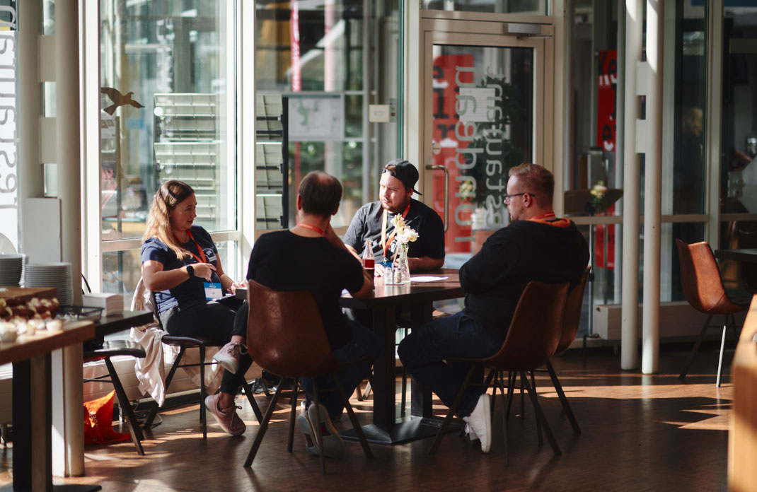Event Foto vom BAR-CAMP 2022 der KELLERKINDER: Vier Teilnehmende sitzen an einem Tisch im sonnendurchfluteten Foyer, vertieft in ein Gespräch. Auf dem Tisch stehen Glasflaschen und ein kleiner Blumenschmuck.