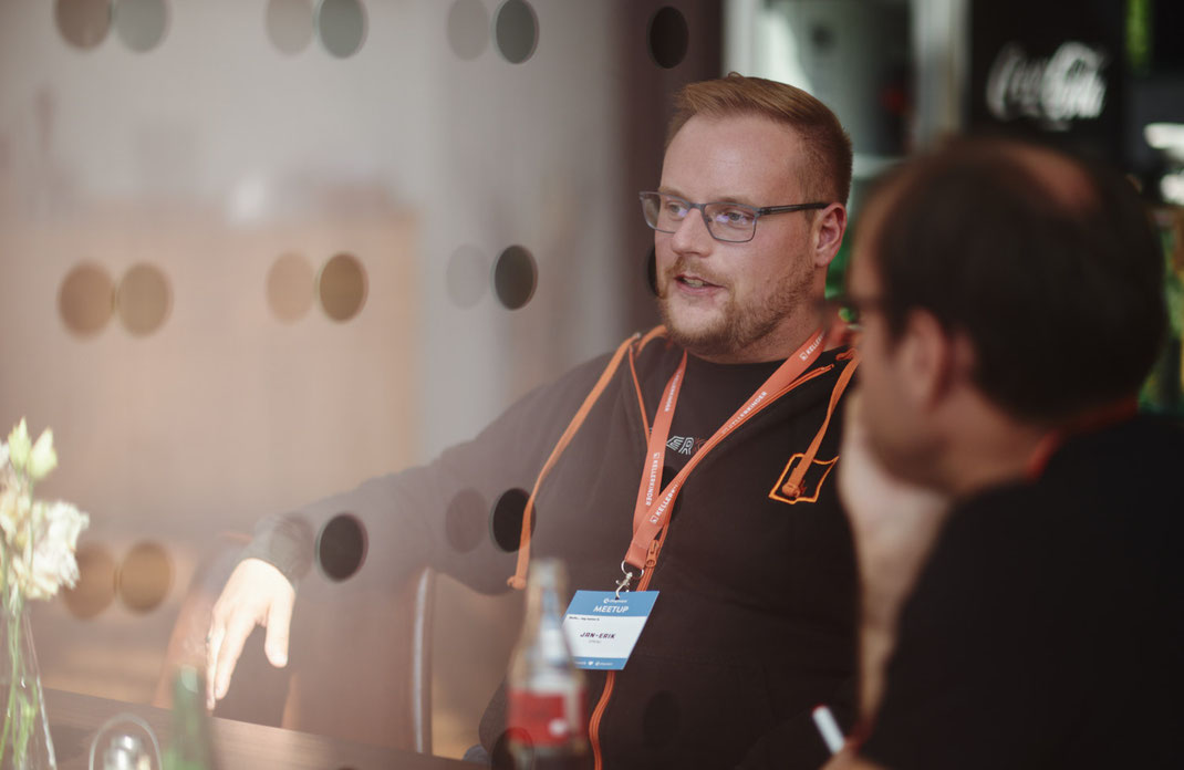 Event Foto vom BAR-CAMP 2022 der KELLERKINDER: Ein Mann mit Brille und Namensschild sitzt an einem Tisch hinter einer Glaswand mit Punktmuster und unterhält sich konzentriert mit einer weiteren Person. Im Vordergrund stehen Flaschen und ein Blumenstrauß.