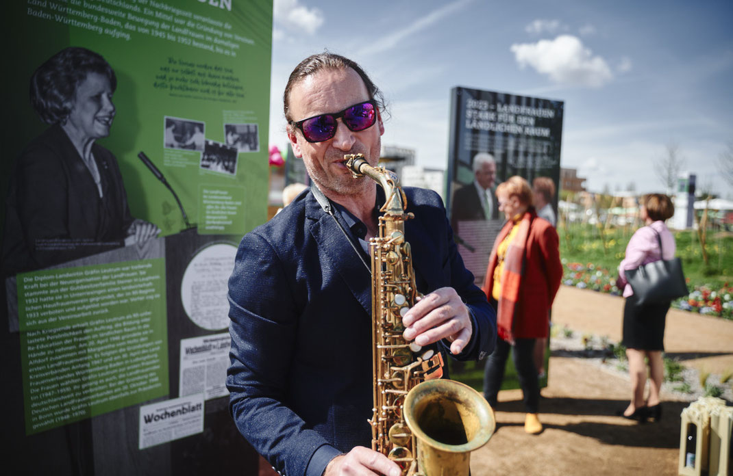 Eventfoto von Christoph Bastert: Saxophonist spielt live bei der LandFrauen-Veranstaltung auf der BUGA Mannheim 2023, mit Ausstellungstafeln im Hintergrund.