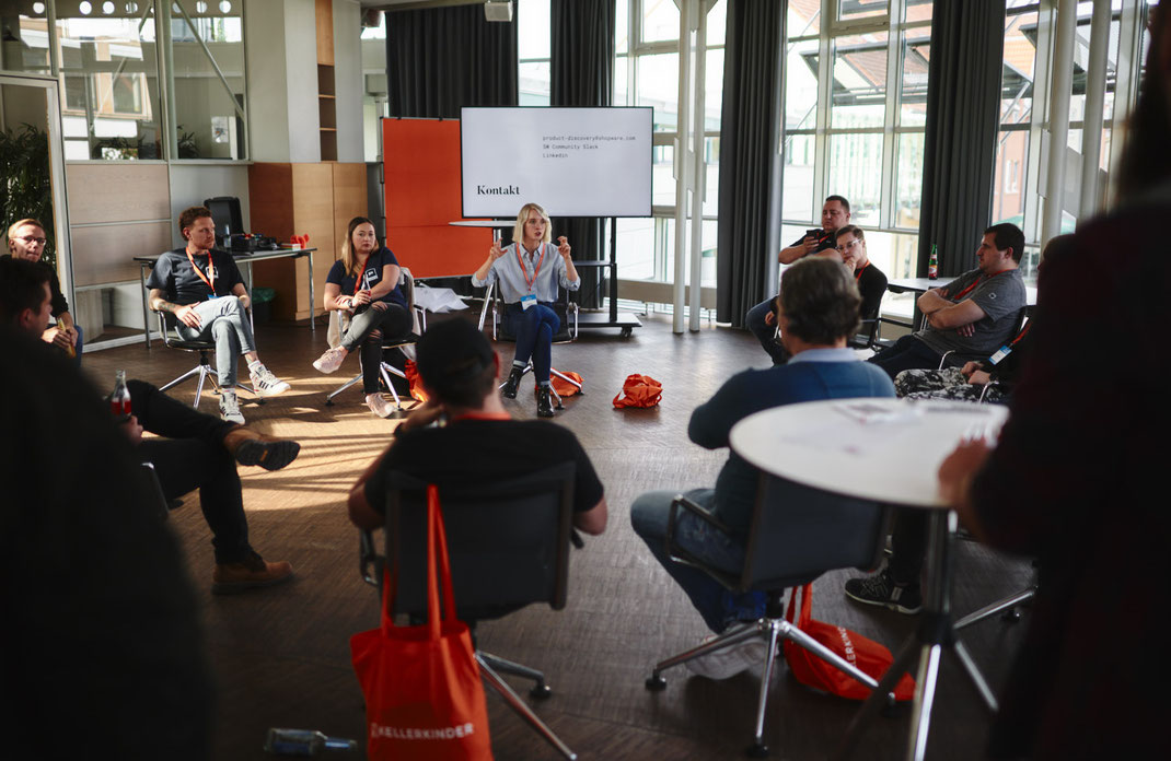 Event Foto vom BAR-CAMP 2022: Teilnehmerinnen und Teilnehmer sitzen in einem Stuhlkreis, eine Frau spricht gestikulierend vor einem großen Bildschirm mit Kontaktinformationen, im Hintergrund ist viel Tageslicht.