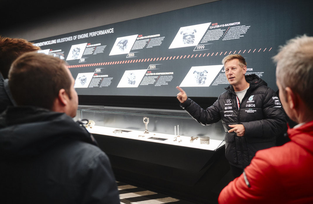 Event Fotos mit technischem Tiefgang Mehrere Personen stehen vor einer Wand mit technischen Abbildungen von Motoren und Daten. Ein Mann in schwarzer AMG-Jacke spricht und gestikuliert, während die Gruppe aufmerksam zuhört. 