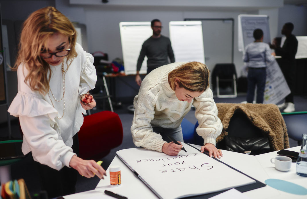 Event-Foto vom Mercedes-Benz Global Training in Stuttgart: Zwei Teilnehmerinnen gestalten gemeinsam ein Flipchart mit der Überschrift „Share your experience“.