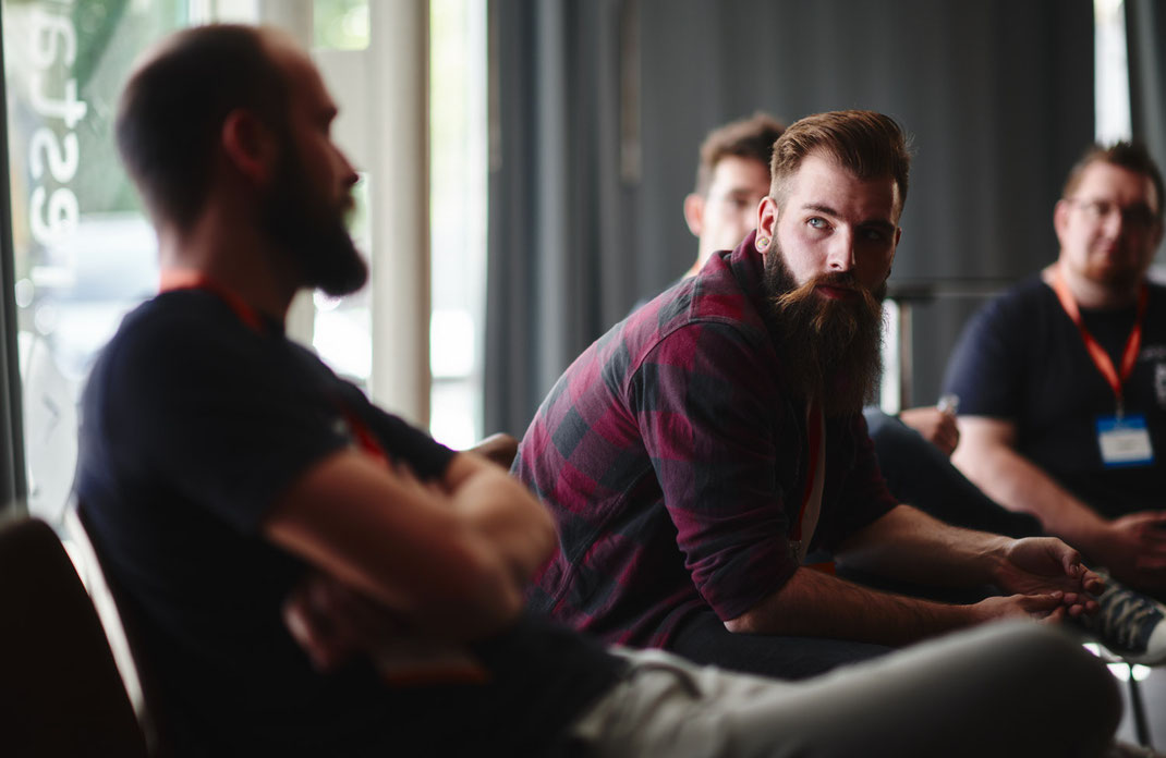 Event Foto vom BAR-CAMP 2022 der KELLERKINDER: Ein Teilnehmer mit Vollbart und Tunnel-Piercings sitzt im Gespräch mit anderen, Licht fällt durch große Fenster auf die Gruppe.