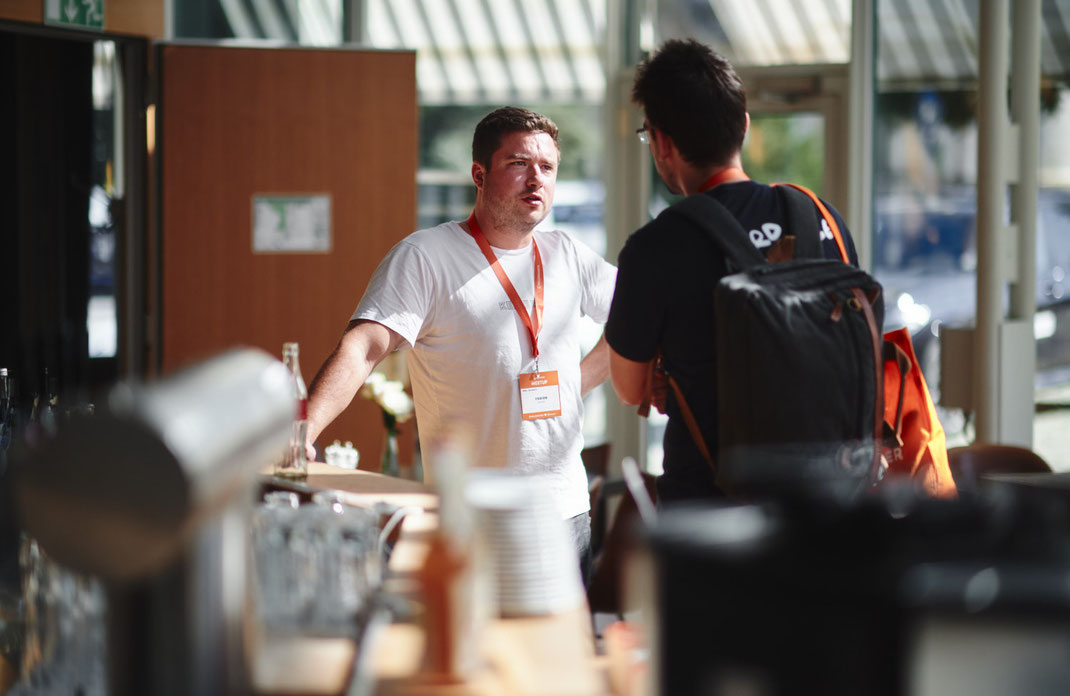Event Foto vom BAR-CAMP 2022 der KELLERKINDER: Zwei Männer stehen an einem Bartresen im Gespräch. Einer trägt ein weißes T-Shirt mit Namensschild, der andere einen Rucksack.
