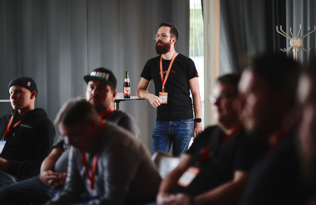 Event-Fotograf beim BAR-CAMP der KELLERKINDER: Ein Teilnehmer mit Bart und Brille lehnt entspannt an einem Stehtisch, im Hintergrund eine Flasche. Im Vordergrund sitzen konzentrierte Zuhörer mit Namensschildern