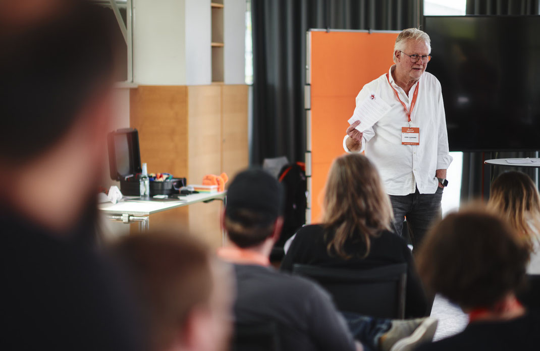 Event Foto vom BAR-CAMP 2022 der KELLERKINDER: Ein Mann mit weißem Hemd spricht vor einem sitzenden Publikum, in der Hand hält er ein paar Zettel.