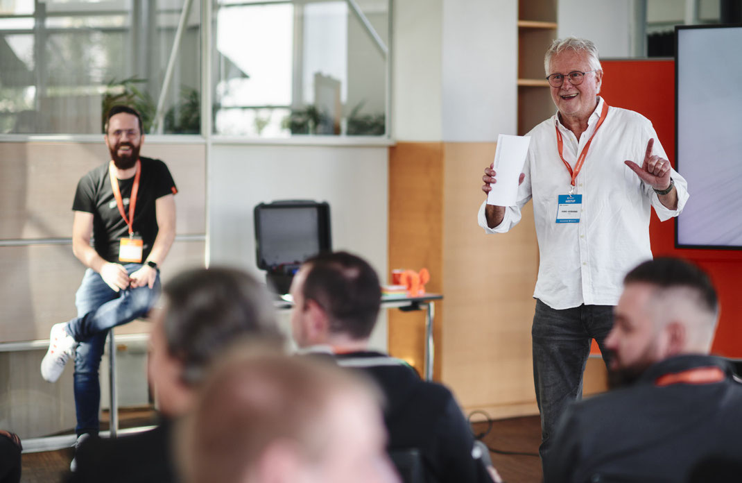 Event Foto vom BAR-CAMP 2022 der KELLERKINDER: Ein Mann steht mit einem Zettel in der Hand vor einer Gruppe Teilnehmender, spricht und gestikuliert. Im Hintergrund sitzt ein weiterer Mann auf einem Barhocker und lacht.