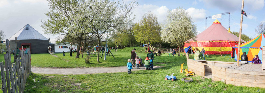 Panoramabild von der Wilden Warte. Zu sehen sind u.a  links im Hintergrund die Zirkushalle, rechts das große Zirkuszelt und daneben ein kleines Zirkuszelt, davor ein Holzschiff zum klettern. Auf dem Platz laufen und stehen einige Personen