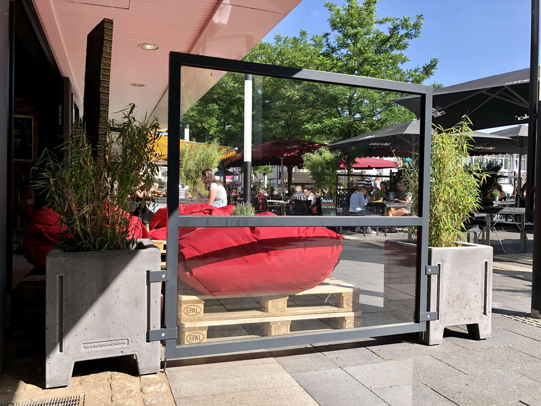 Mobiles Windschutzelement aus Glas und Aluminium, eingefasst in Beton-Pflanzkübel an einem Outdoor-Gastronomiebereich