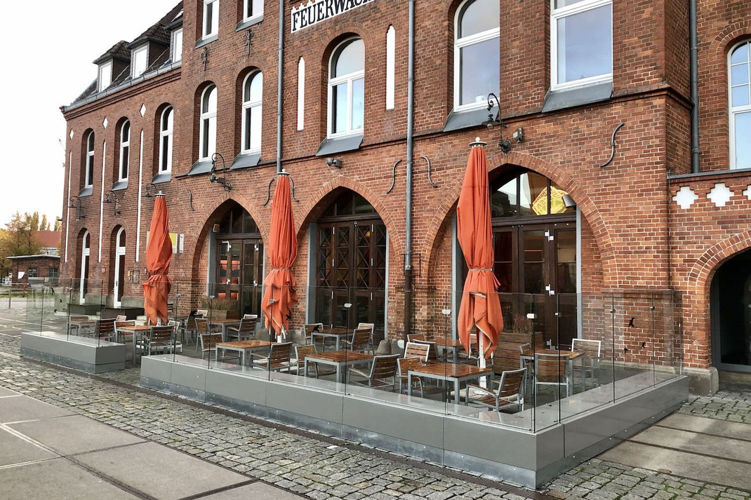 Windschutz-Anlage mit Glas-Elementen vor einem historischen Backsteingebäude, ergänzt durch Sonnenschirme auf einer Restaurant-Terrasse