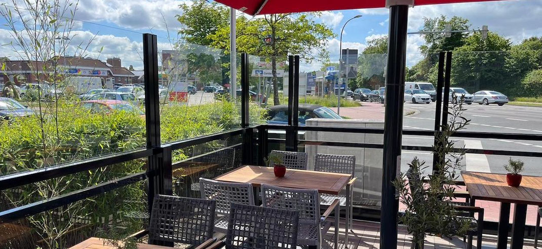 Biergarten mit einem in der Höhe verstellbaren Windschutz aus Glas und Aluminium an einer Straße als Lärmschutz
