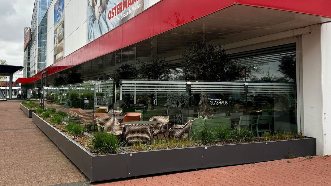 Windschutz-Anlage als Abgrenzung der Terrasse für die Außengastronomie eines Möbelhauses bestehend aus anthrazitfarbenen Sockelmodulen  transparenten Glas-Elementen.