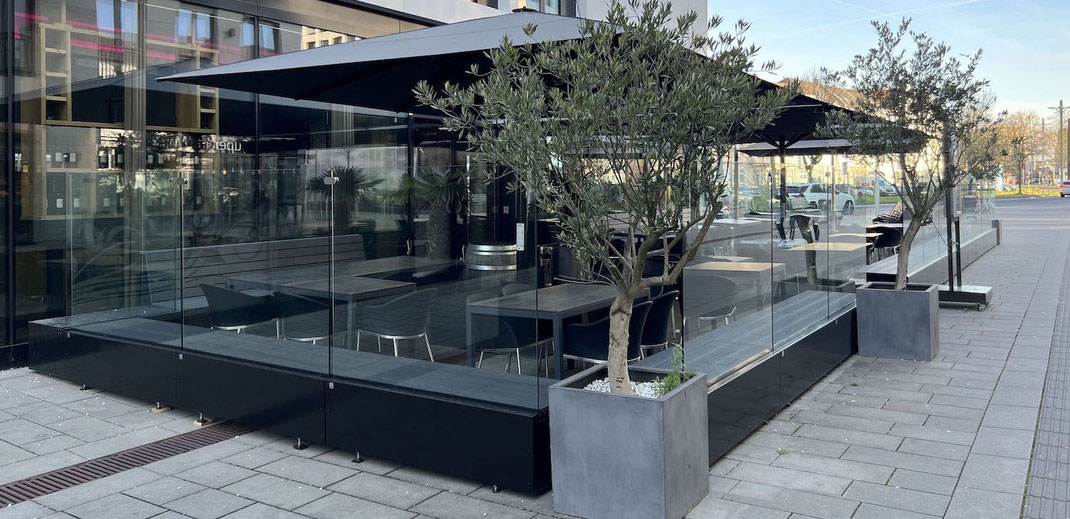 Moderne Terrassengestaltung eines Restaurants mit transparentem Glas-Windschutzund großen schwarzen Sonnenschirmen für eine windgeschützte Außengastronomie