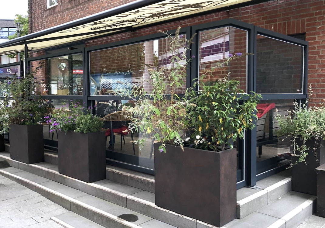 höhenverstellbaren Windschutzes Modell Athen – passgenauer Wetterschutz auf einer Gastronomie-Terrasse in Kombination mit dekorativen Pflanzgefäßen