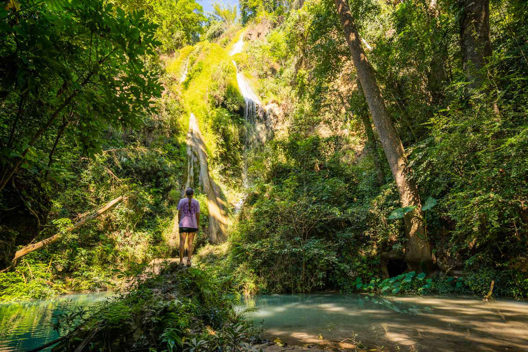 Una mujer con una camiseta morada está de pie junto a un charco de agua con vistas a la cascada.