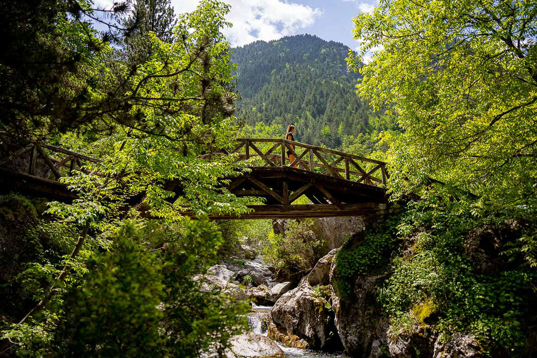 Frau geht auf einer Holzbrücke über einen reißenden Bergbach, im Hintergrund sieht man die bewaldeten Berghänge.