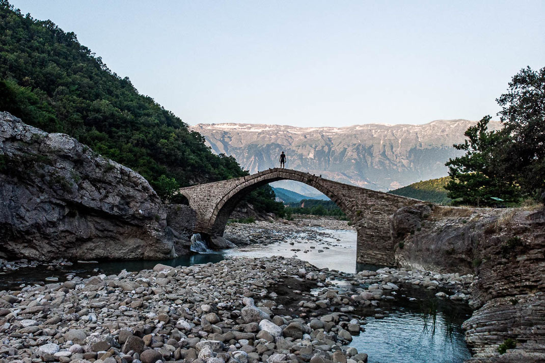 Mann steht auf Brücke bei Heißen Quellen über einem Fluss mit Blick in Richtung Berge.