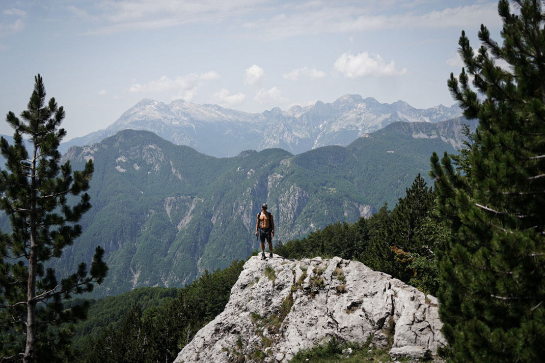 Durchtrainierter Mann steht auf Berggipfel, im Hintergrund sieht man die bewaldeten Berge.