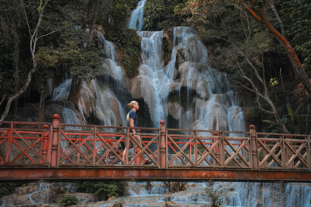 A man walks on a red bridge and looks at Tat Kuang Si waterfall.