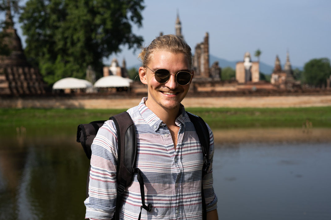 Un homme sourit à la caméra avec les ruines de la ville de Sukhothai en arrière-plan.