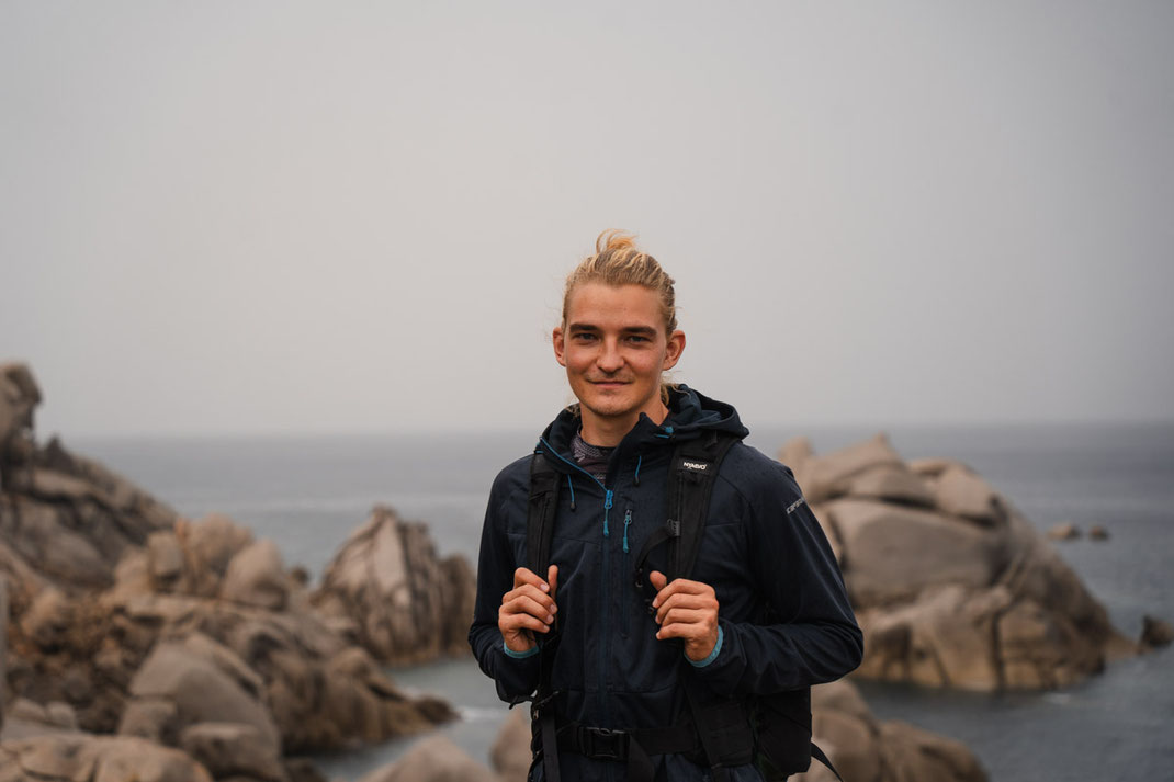 Ein Mann blickt in die Kamera, im Hintergrund die Felsen des Capo Testa.