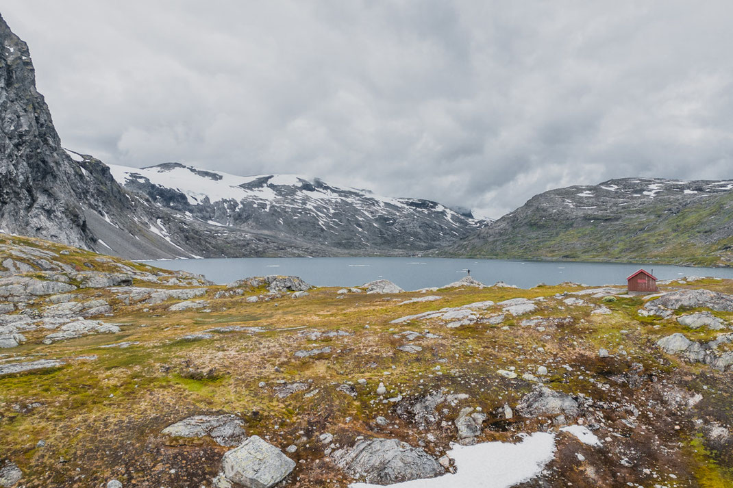 Ein Mann steht auf einem Felsen vor einem Gletschersee in der beeindruckenden Landschaft Norwegens, auf der rechten Seiten steht ein rotes Haus.