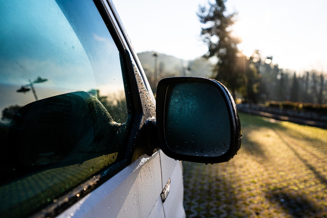 Seitenspiegel eines weißen VW-Busses mit gefrorener Scheibe während Sonnenaufgang.
