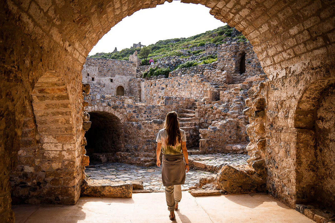 Frau geht in Monemvasia durch einen malerischen Torbogen, im Hintergrund sind die Ruinen einer Burg zu sehen.
