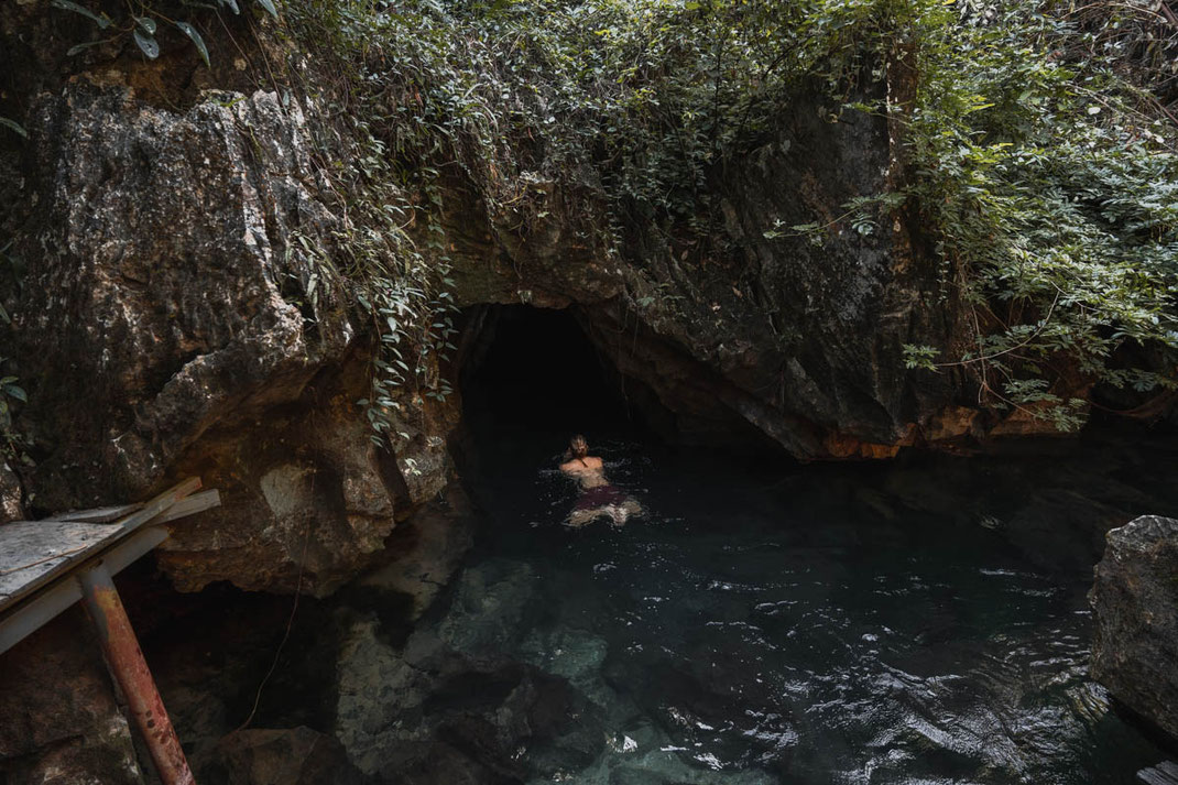 Un hombre nada en una pequeña laguna hacia una cueva adyacente.