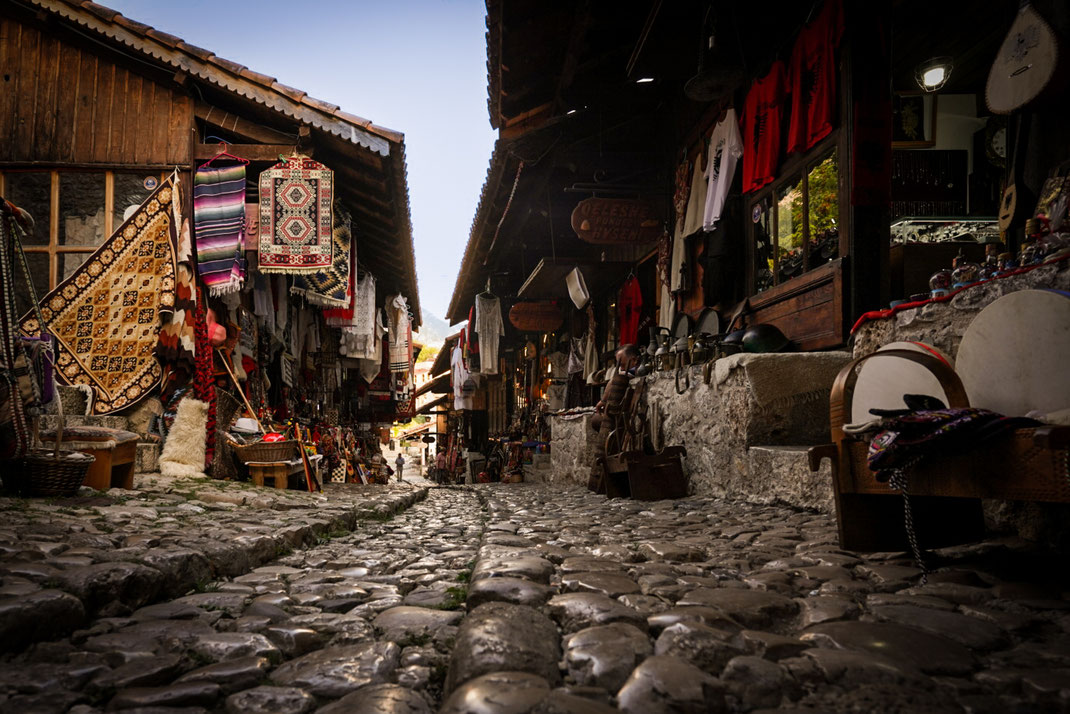 Malerische Marktgasse im alten Basar von Kruja mit gepflastertem Weg und kleinen Geschäften, die Teppiche, traditionelle Kleidungsstücke und Souvenirs anbiete.