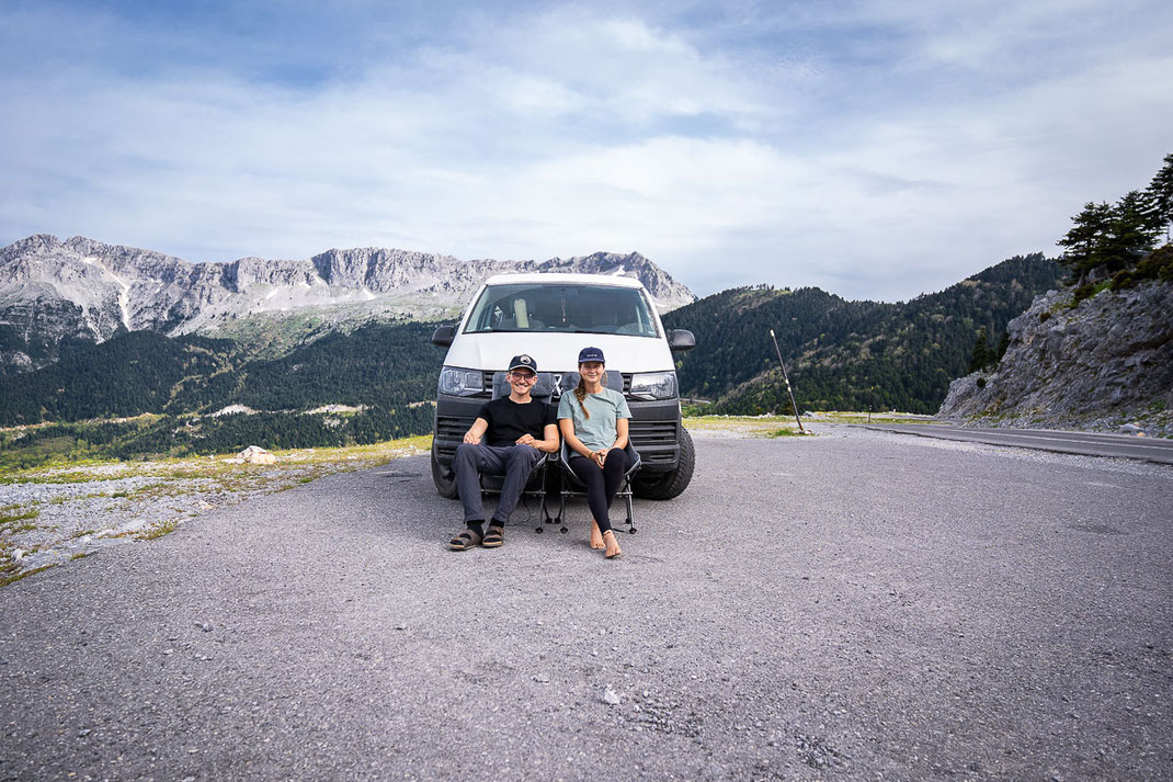 Ein Pärchen sitzt vor ihren VW-Bus und lächelt in die Kamera, im Hintergrund sind hohe Berge.