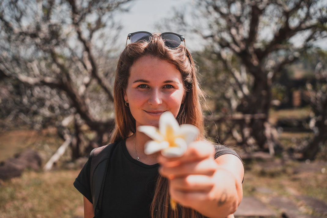 Une femme tient une fleur blanche devant la caméra en souriant.