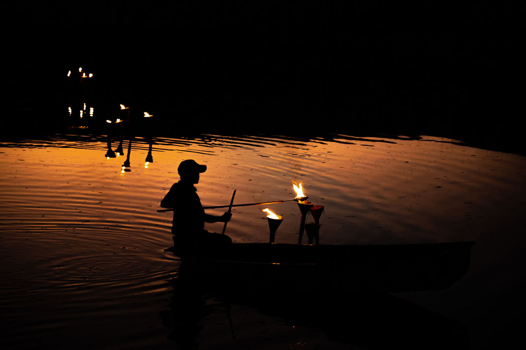 A man in a small boat lights the torches in the water.