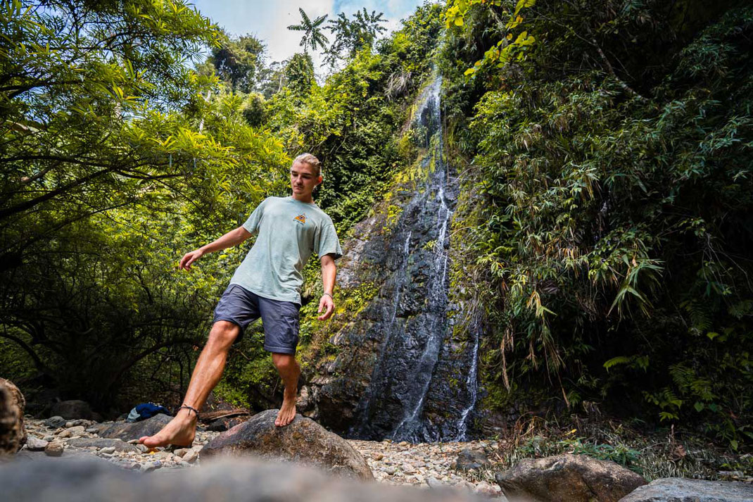 Ein Mann balanciert über Steine mit dem Kaeng Nyui Wasserfall im Hintergrund.