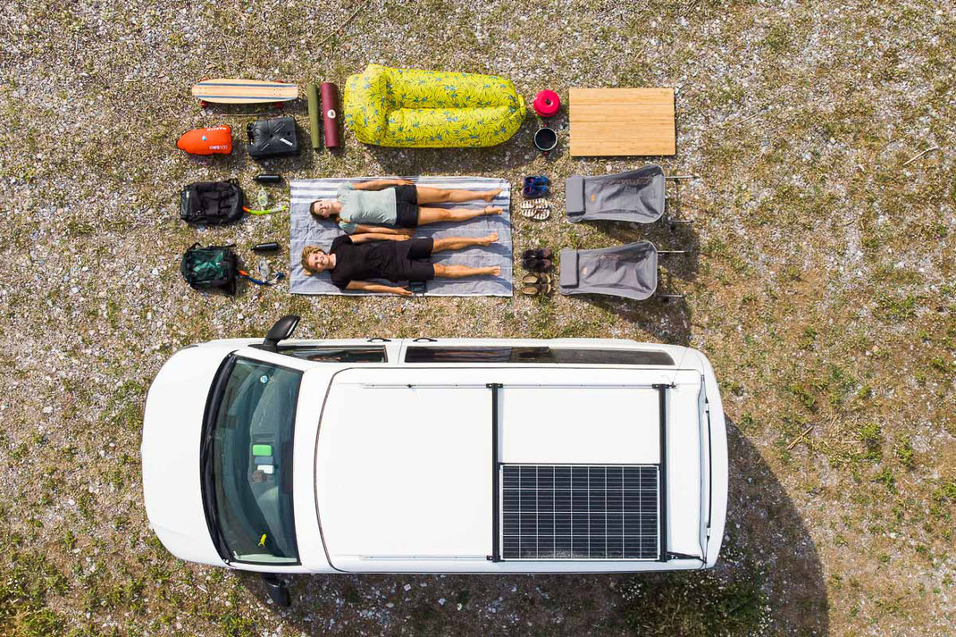 Drohnenaufnahme von weißem VW Bus mit Solaranlage und jungem Pärchen, die mit ihrem Gepäckstücken am Boden liegen.
