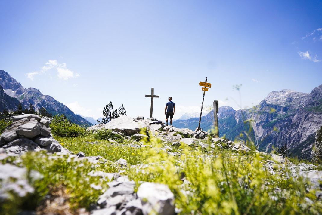 Mann steht am Gipfelkreuz und genießt Aussicht am Peja-Pass auf das Tal von Theth.