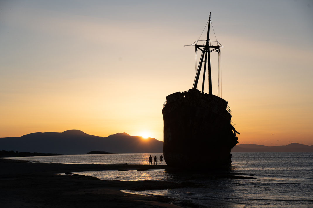 Menschen stehen vor dem Schiffswrack am Meer und beobachten den Sonnenaufgang.