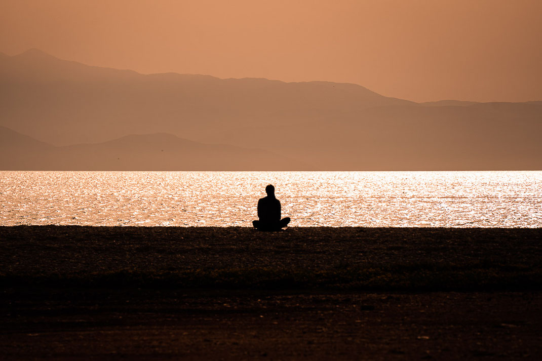 Eine Frau sitzt am Strand bei Sonnenuntergang und blickt auf das Meer hinaus.