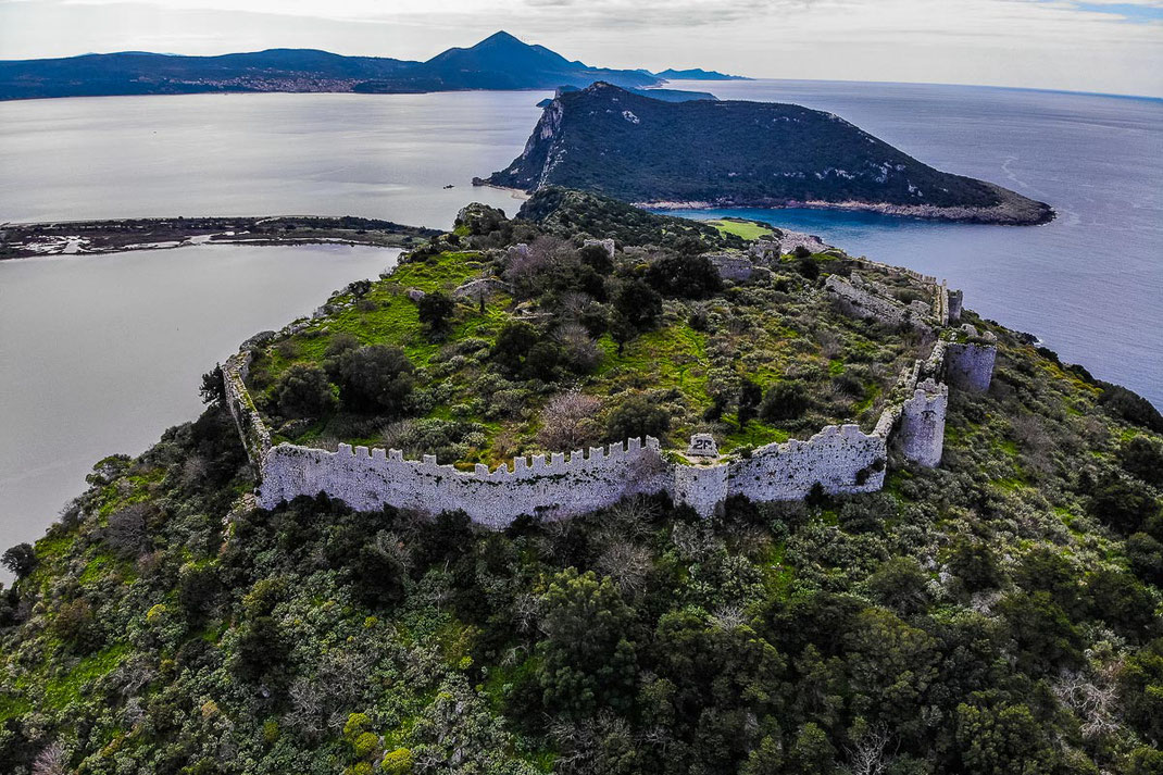 Das von grünen Wiesen und Bäumen überzogene Paliokastro, auch Nestors Schloss genannt, aus der Vogelperspektive mit Meer zur Rechten und Salzwasserlagune zur Linken.